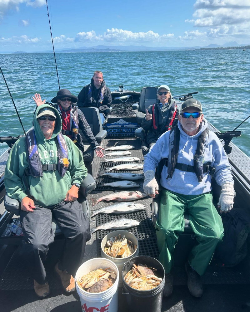 A family of four posing happily on a fishing boat with eight large salmon lined up on the diamond plate deck under a cloudy sky.