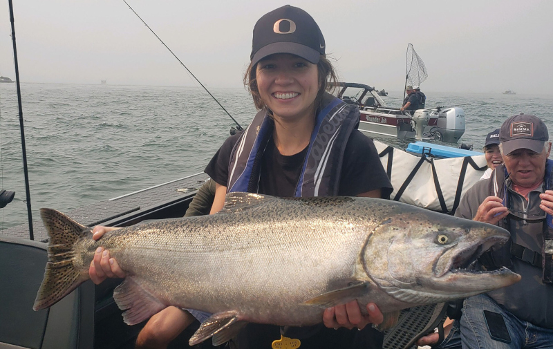 A smiling female angler on a boat holding a massive silver Chinook salmon caught during a peak season charter on the Oregon coast.