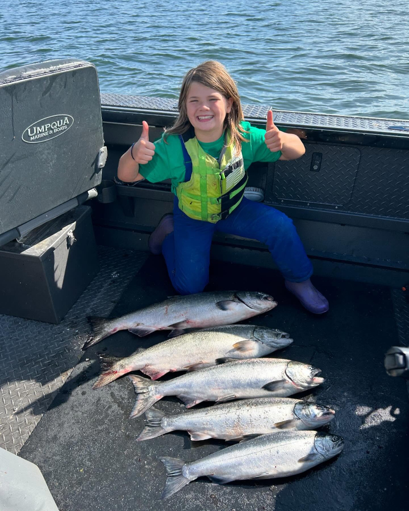 A young girl in a life jacket giving two thumbs up while kneeling behind five freshly caught silver salmon on the deck of a boat.